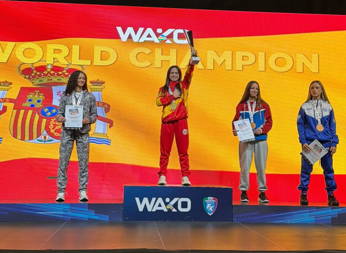 Andrea Martínez Terres brandit le trophée sur la plus haute marche du podium du Championnat du monde WAKO à Jesolo, devant le drapeau espagnol affiché à l'écran